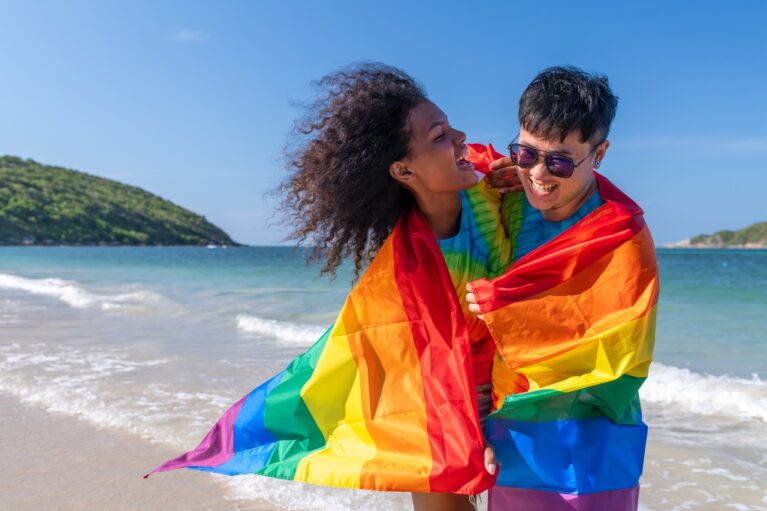 Two people smiling and wrapped in a rainbow pride flag stand on a sunny, gay-friendly Curaçao beach with gentle waves, green hills, and a blue sky in the background.
